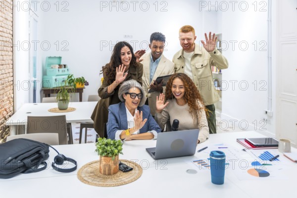 Diverse business colleagues smiling and waving at a laptop during a video call, connecting with remote team members in a modern office for virtual collaboration and teamwork