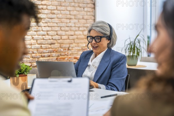 Senior businesswoman with a headset providing attentive customer service and digital support while sitting at a desk with a laptop, assisting clients in a modern office