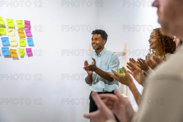 Smiling business team members are clapping, showing appreciation and recognition during a successful office meeting or presentation, with colorful sticky notes on a wall