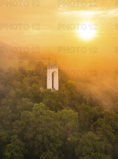 White tower rises out of misty forest while the sun illuminates orange sky, Schönbergturm, Pfullingen, Swabian Alb, Germany