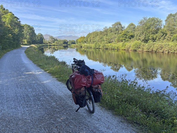 A loaded bike ride along a river under clear skies and lush vegetation, Caledonian Canal, Fort William, Scotland, United Kingdom