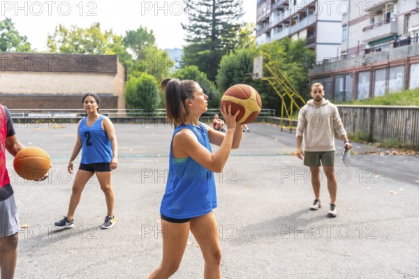 Concentrated female basketball player in blue uniform shooting ball during training outdoors on a sunny day, watched by coach with clipboard and teammates