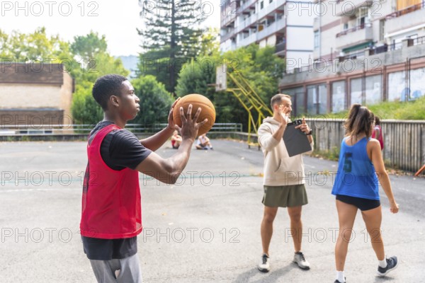 Young basketball player practicing shooting technique on outdoor court, receiving guidance from coach who's holding clipboard and giving instructions while other players observe