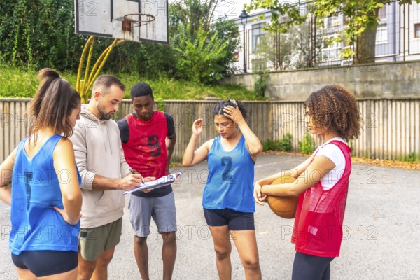Coach writing notes on a clipboard while providing instructions to male and female basketball players during an outdoor training session on the court, fostering teamwork and skill development