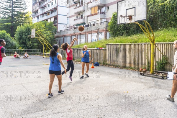 Female basketball player jumping and shooting the ball during a training session on an outdoor court, with her teammates and coach watching