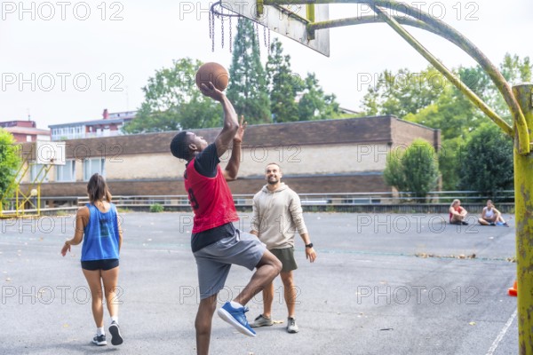 Group of friends enjoying a game of basketball on an outdoor court, with a young man shooting the ball while his friends cheer him on, embodying teamwork and camaraderie