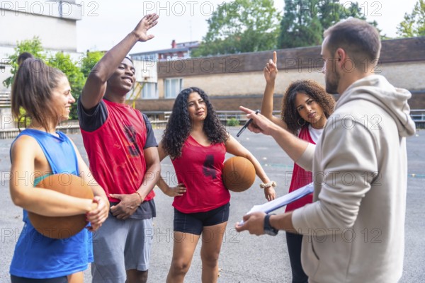 Basketball coach holding a clipboard, explaining game strategy while engaging with team players during an outdoor training session on the court, fostering teamwork and skill development