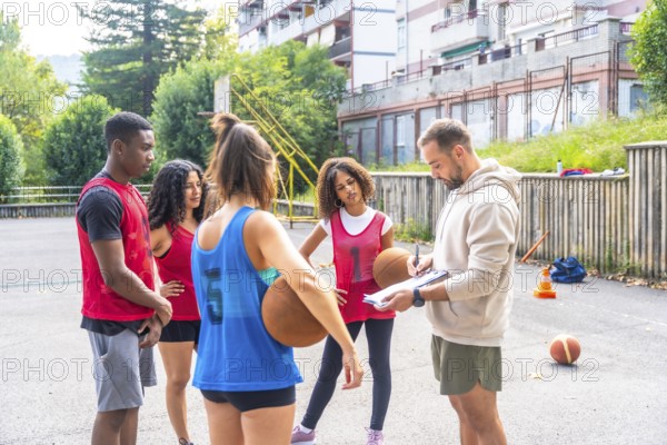 Basketball coach writing notes on clipboard while giving instructions to multi ethnic team players during training session on outdoor court