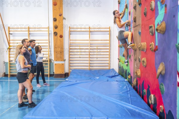 Determined sportswoman practicing bouldering in a climbing gym, ascending a vibrant wall as friends watch and support her, highlighting strength, skill, and camaraderie