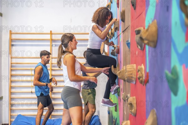 Young woman is practicing sport climbing on a colorful climbing wall in a gym, her instructor is supporting her while other athletes train in the background