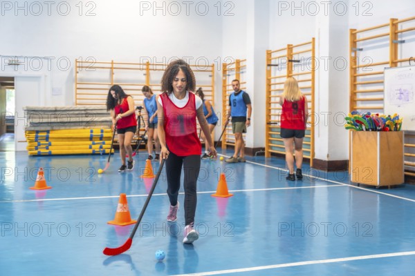 Young woman practicing floor hockey, dribbling the ball with her stick, during a training session in a gym, surrounded by teammates and coaches, focused on improving her skills and teamwork