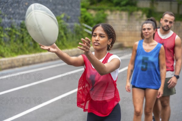 Young woman wearing a red training bib practices passing a rugby ball on a sports field, while other players watch in the background, during a training session