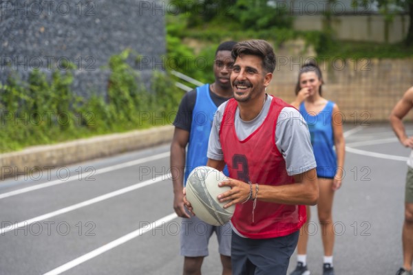 Rugby player wearing a red training bib smiling while holding the ball during a lively training session with teammates on an outdoor court, embodying teamwork and athletic spirit