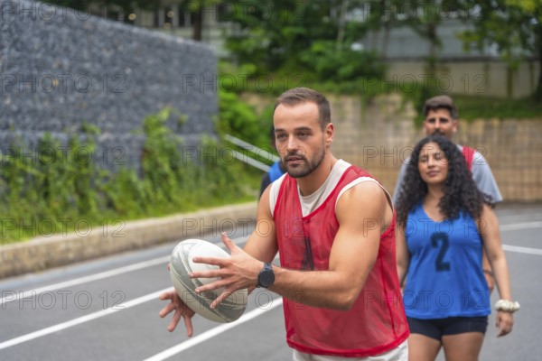 Male rugby player wearing red training bib holding and passing rugby ball on outdoor court during team practice, with teammates walking behind him
