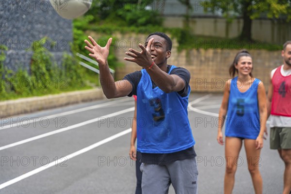 Young male athlete wearing a blue training bib catching a rugby ball during practice, with his teammates watching and smiling in the background on an outdoor court