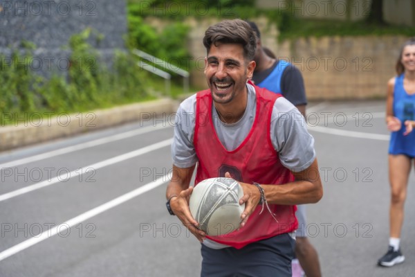 Male rugby player wearing a red training bib smiles broadly while holding the ball during practice on an outdoor court, with other players in the background