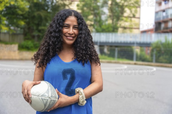 Young woman wearing a blue rugby uniform, confidently holding a rugby ball while smiling on an outdoor court, showcasing readiness for the upcoming game and embracing the spirit of competition