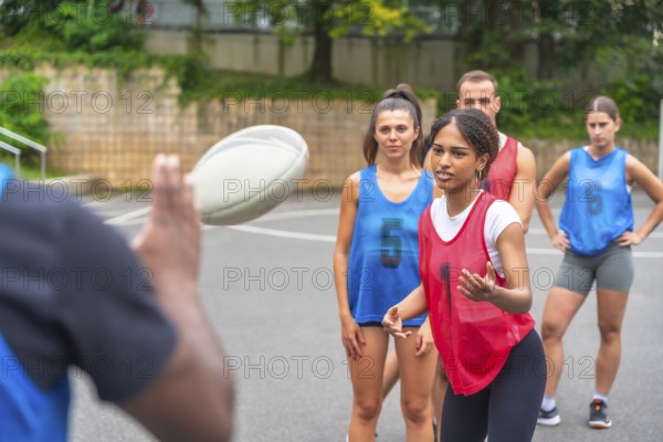 Coach throws a rugby ball to a group of female players wearing numbered jerseys during a training session on an outdoor court, fostering teamwork and athletic skills