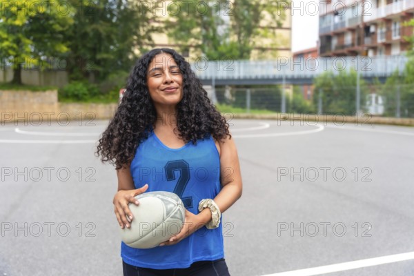 Female rugby player wearing a blue shirt, smiling confidently while holding a rugby ball on an urban court, poised for training or an upcoming match