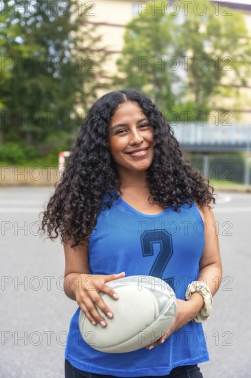 Confident female rugby player is smiling while holding a rugby ball, standing on a sports field, ready for the game, showcasing strength and determination
