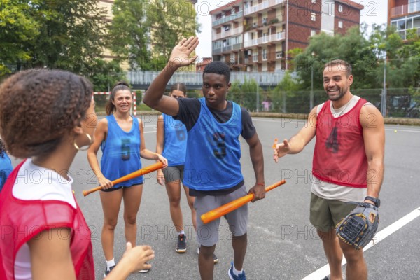 Group of cheerful multi ethnic friends wearing sportswear giving high fives and holding bats while enjoying a friendly baseball match on a sunny day on an outdoor court