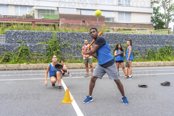 Baseball player swings the bat, preparing to hit the ball during a training session, while teammates watch and wait their turn on a city field