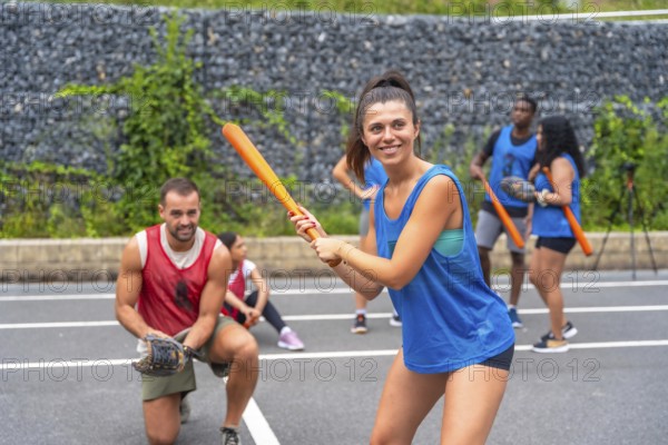 Smiling sportswoman holding an orange baseball bat, getting ready to hit the ball during a friendly baseball game with friends on an outdoor court