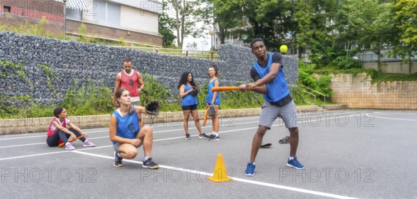 Multi ethnic baseball players practicing on an urban field, with a batter hitting the ball and a catcher poised to receive, highlighting teamwork and athleticism