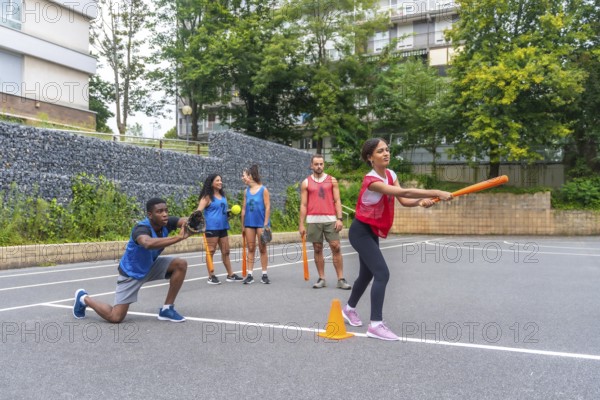 Group of friends enjoying a lively game of baseball on an outdoor court, with a woman swinging an orange bat while a male friend gets ready to catch the ball