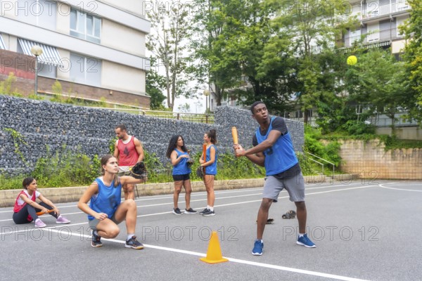 Group of young friends playing baseball in an urban environment, with a male player hitting the ball while his teammates watch and wait their turn