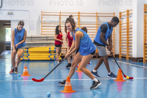 Female and male athletes wearing blue t shirts are practicing floorball, dribbling with sticks around orange cones in a gym with a blue floor and yellow equipment