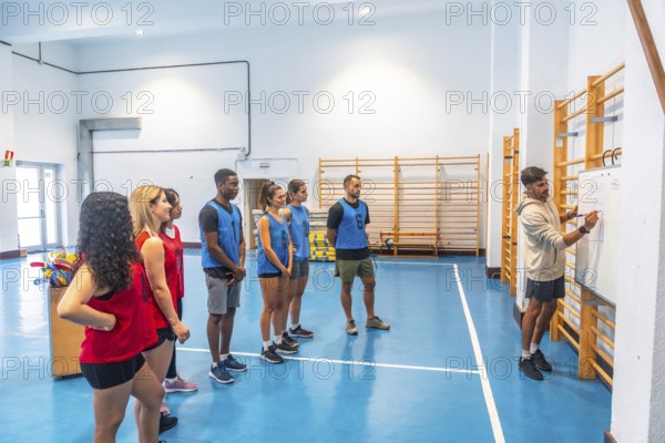 Hockey coach drawing a game plan on whiteboard while explaining strategy to players divided in two teams, wearing red and blue uniforms, listening attentively in a gymnasium