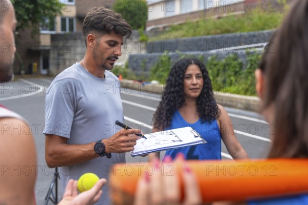Baseball coach drawing a play on a clipboard while explaining game strategy to his team on an outdoor court, fostering teamwork and concentration during practice discussions
