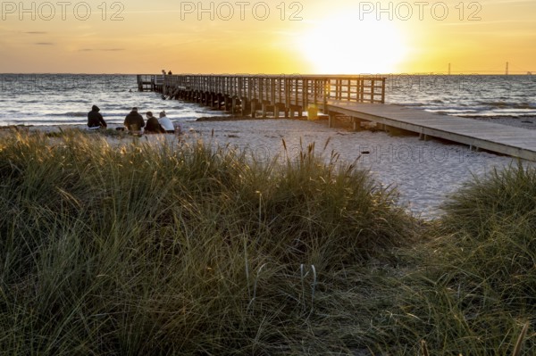 People sitting on the beach while a long pier leads into the sea, framed by sunset lights, Kobaek Strand, Sjaelland, Denmark