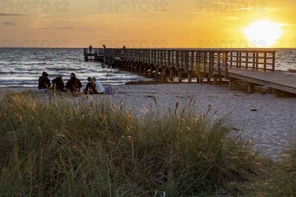 Group of people sitting on the beach next to a pier at sunset, surrounded by grass and sea, Kobaek Strand, Sjaelland, Denmark