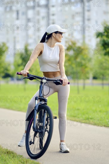 A slender woman wearing activewear and a cap takes a break from cycling. She stands beside her bike on a path lined with greenery and modern buildings, enjoying the beautiful weather