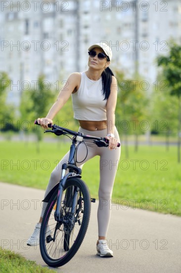A woman sits on her bicycle in a vibrant park, dressed in a light sports outfit. She appears relaxed, enjoying the sunny weather amidst green trees and modern buildings in the background