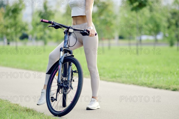 Slender female legs in athletic wear stand beside the mountain bike on a smooth pathway in a park. Green trees and grass are visible in the background, creating a serene atmosphere