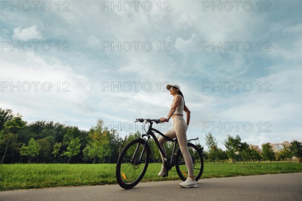 A woman stands beside her bicycle in a lush green park. She wears comfortable athletic clothing and a cap, the sky is partly cloudy, enhancing the calm setting
