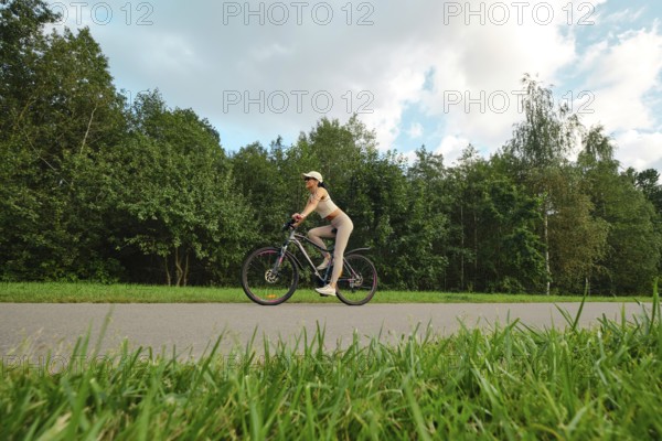 A woman in athletic wear rides her bicycle on a smooth path surrounded by lush green trees and grass. The bright sky adds to the serene outdoor atmosphere during a pleasant day