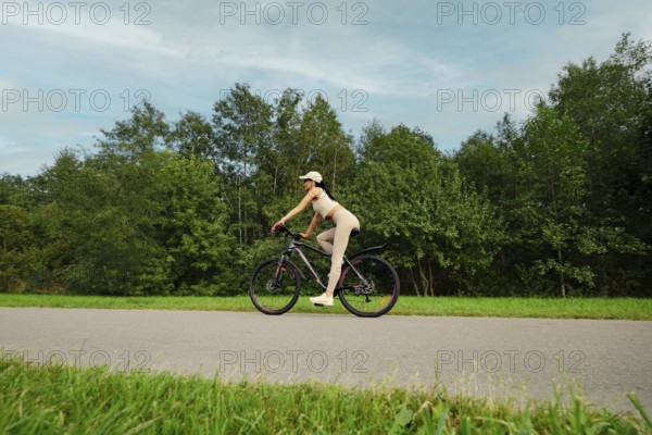 A woman rides her mountain bike on a smooth path in a lush green landscape. Tall trees line both sides of the trail, creating a tranquil atmosphere during a bright day