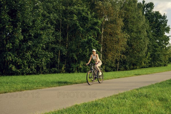 A woman rides a bicycle along a paved path surrounded by tall trees and vibrant greenery. The sunny weather creates a cheerful atmosphere for outdoor activities
