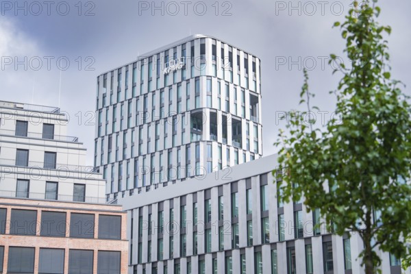 Modern high-rise building with glass façade under a cloudy sky, bordered by a tree, Munich, Germany