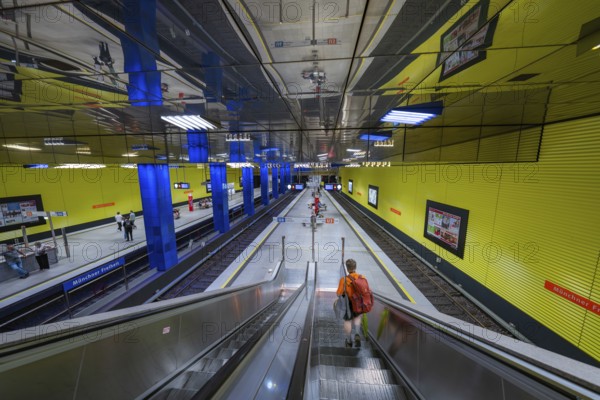 Modern underground station with yellow walls and escalator, people in the hall, Münchner Freiheit, Munich, Germany