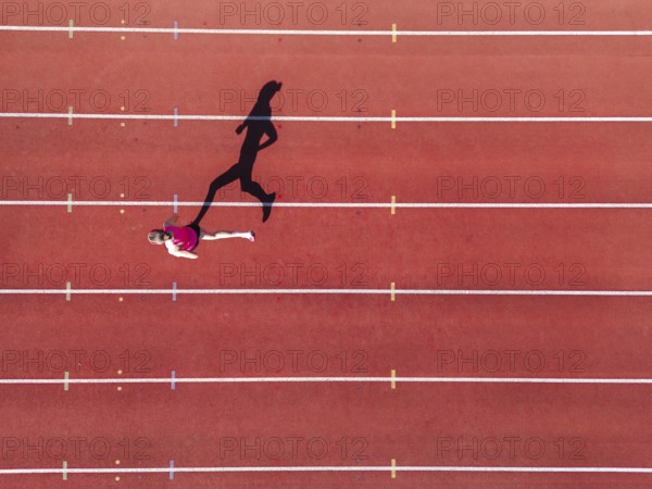 Aerial view of a female individual (runner) on a red athletics track (tartan track) with shadow as a silhouette