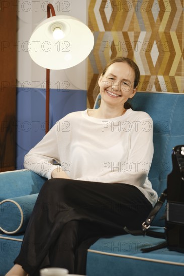 A woman sits comfortably on a vibrant blue couch, smiling while relaxing in a stylish living room featuring warm lighting and geometric decor. The atmosphere exudes a sense of calm and comfort