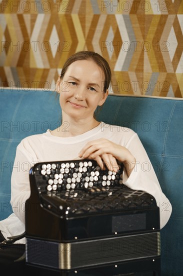 A middle-aged woman in a casual white top smiles, resting her hands on an accordion in a vibrant, geometric-patterned room, capturing a joyful moment of music and creativity