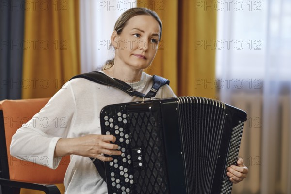 A woman sits comfortably in a chair, skillfully playing the accordion in a well-lit room adorned with warm curtains. The atmosphere is relaxed, perfect for a musical gathering or practice
