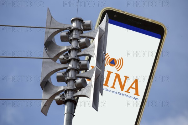 (Photomontage) Close-up of a siren, next to it the open warning app NINA on a smartphone