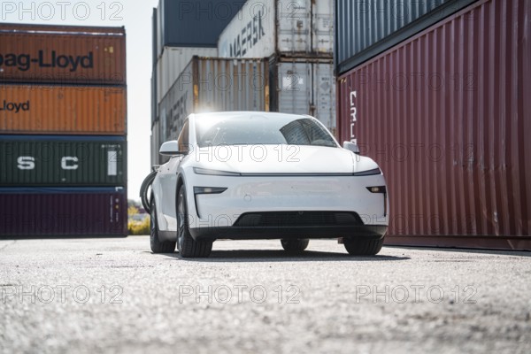 White car in a harbour environment next to stacked freight containers, Tesla Model Y Juniper, electric car